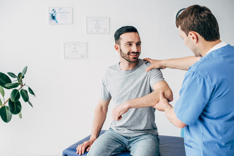 Smiling patient sitting on couch and doctor examining patient shoulder in massage cabinet at clinic